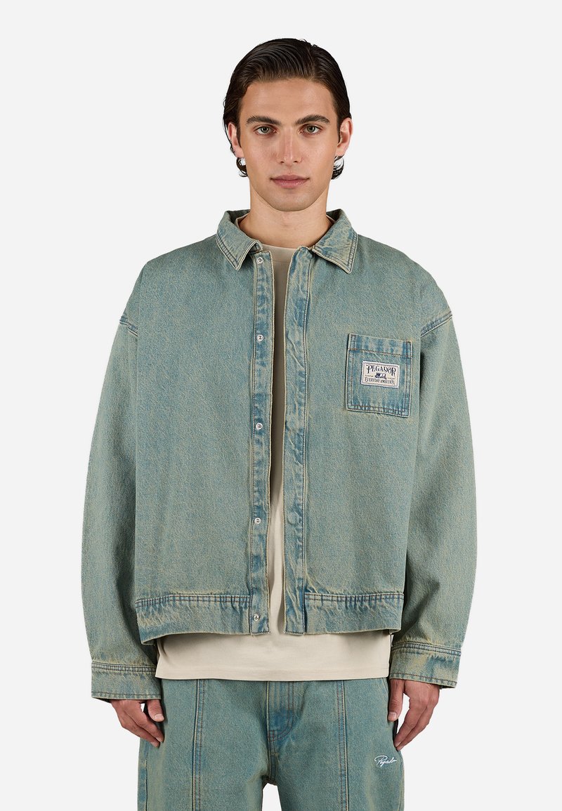 Chaqueta de denim azul claro con forma cuadrada, bolsillo frontal y cierre de botones. Presenta una textura desgastada y detalles de costura visibles.