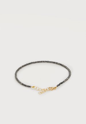 Bracelet délicat avec de petites perles foncées, fermoir doré et chaînette d’extension, posé à plat sur un fond clair.