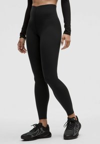 Sorte højtaljede leggings lavet af strækbart materiale med en glat overflade. Parret med sorte træningssko med netdetaljer.