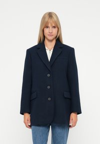 Donna con capelli biondi lisci che indossa un blazer oversize blu scuro sopra una maglietta bianca con zip e jeans blu, su uno sfondo bianco.
