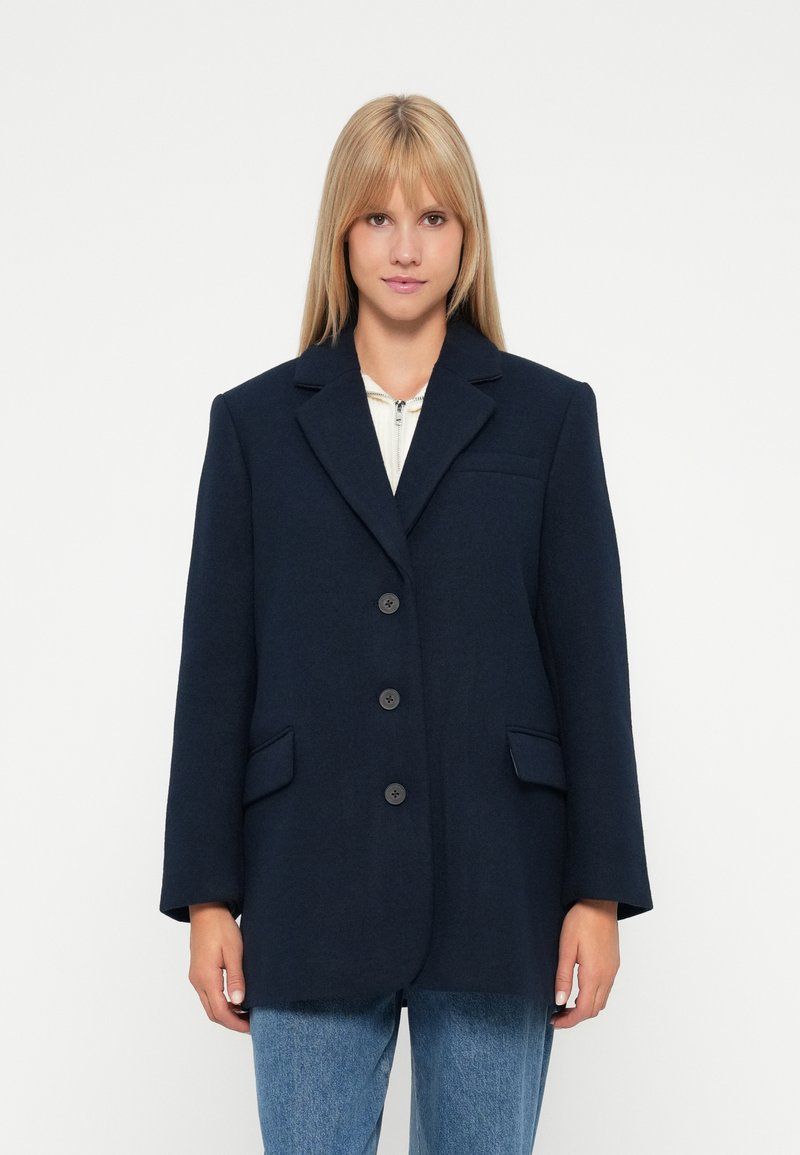 Donna con capelli biondi lisci che indossa un blazer oversize blu scuro sopra una maglietta bianca con zip e jeans blu, su uno sfondo bianco.