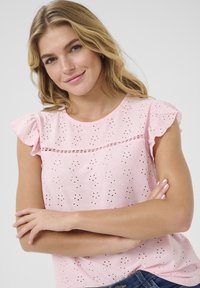 Lys pink kortærmet top med flæser på ærmerne og hulmønster. Pyntet med små dekorative elementer under halsudskæringen. Blødt og åndbart stof.