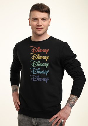 Mężczyzna o krótkich brązowych włosach, z tatuażami na przedramionach, ubrany w czarną bluzę z napisem "Disney" w pięciu różnych kolorach tęczy.