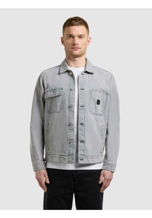 Mann trägt eine hellgraue Jacke mit Knöpfen und zwei Brusttaschen über einem weißen Hemd und schwarzen Hosen, steht vor einfarbigem Hintergrund.