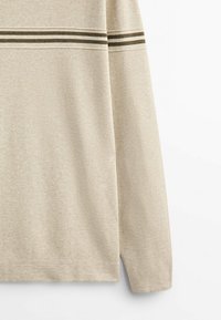 Beżowa koszula z długim rękawem o teksturowanym wzorze, z dwoma poziomymi oliwkowo-zielonymi pasekami na klatce piersiowej. Luźny krój i okrągły dół.