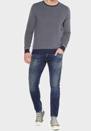 Homme portant un pull à manches longues bleu à motifs, un jean bleu délavé et des baskets montantes grises, debout avec une main dans la poche.