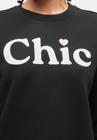 Person wearing a black sweatshirt with white "Chic" text, where the dot on the "i" is replaced by a small pink heart.