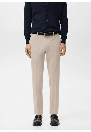 Pantalones beige a medida con corte recto, tejido texturizado y pliegues delanteros, combinados con un cárdigan de botones azul marino y zapatos negros.