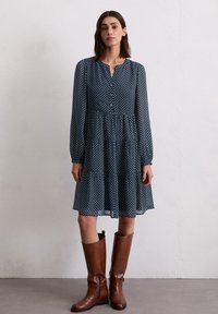 Marineblauwe jurk met een knoopsluiting aan de voorkant, lange mouwen en een gelaagde rok, met een bloemenpatroon; gecombineerd met hoge bruine leren laarzen.