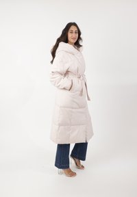 Abrigo acolchado de color rosa claro con capucha, presenta una cintura ceñida con cinturón, bolsillos laterales y un diseño acolchado, combinado con jeans oscuros y tacones transparentes.