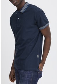 Mørkeblå polo-shirt i bomuld med en struktureret krave og ærmekanter. Har et mærket logo og sidelabel. Bæres med denimjeans.