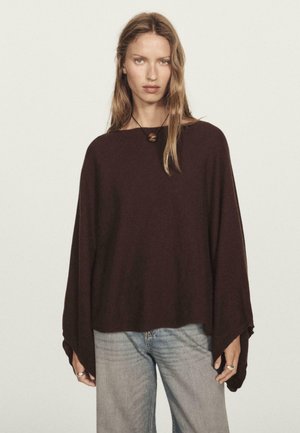 Femme aux cheveux longs et blonds portant un pull poncho ample de couleur brun foncé, un jean bleu clair et un collier avec un pendentif rond, se tenant devant un fond uni.