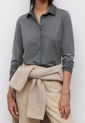 Femme portant une chemise grise à manches longues boutonnée, un pull en maille beige noué autour de la taille et un pantalon beige sur fond blanc.
