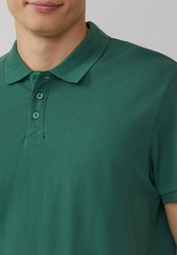 Zielona koszulka polo z teksturowanej tkaniny, z klasycznym kołnierzykiem, trzema guzikami i krótkimi rękawami.