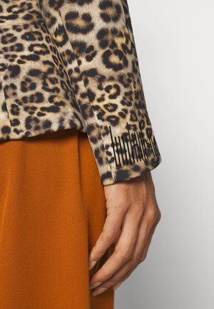 Długi rękaw topu w panterkę z mankietem na tle zapieczonego pomarańczowego, fakturowanego materiału, pokazujący zrelaksowaną dłoń u boku.