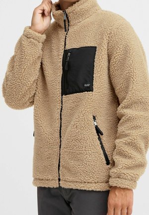 Beige fleecejakke med sort lynlåslomme på brystet og sidelommer, båret af en person, der justerer kraven, mod en ensfarvet baggrund.