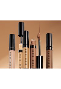 Fünf Bobbi Brown Foundationflaschen in verschiedenen Farbtönen, eine mit offenem Applikator, aus dem flüssige Foundation tropft, vor einem beigen Hintergrund.