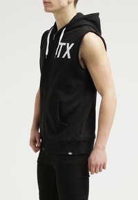 Felpa con zip nera senza maniche, dotata di tasche frontali, lacci bianchi e una grafica bianca "PTX" sul petto. Tessuto morbido.