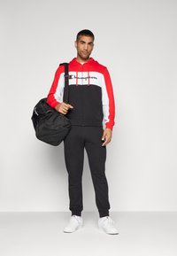Chándal negro con una chaqueta con capucha de cremalleras en rojo y blanco con logo, combinado con pantalones negros y zapatillas blancas, llevando una bolsa de deporte negra.