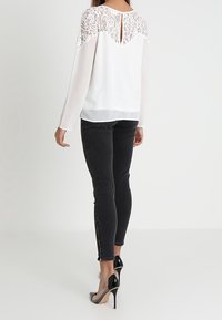 Blusa bianca con dettagli in pizzo sulle spalle e sul retro, e maniche lunghe, abbinata a jeans skinny neri e scarpe con tacco nere.