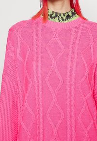 Rosa stickad tröja med kabeldesign och diamantmönster, strukturerade ärmar och ribbad halsringning, över en grön mönstrad topp.