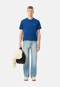 Camiseta de algodón azul de manga corta con un pequeño logo, combinada con unos vaqueros azules desgastados y zapatillas blancas. Sosteniendo un bolso negro y un suéter color crema.