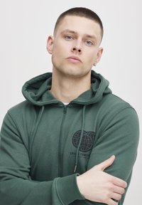Zöld cipzáras kapucnis pulóver, puha anyagból készült, elülső zsebbel és egymásba fonódó köröket ábrázoló logóval. Húzózsinóros kapucnival és bordás mandzsettákkal rendelkezik.