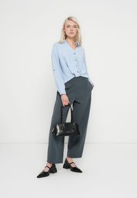 Chemise bleu clair texturée à boutons avec manches longues, associée à un pantalon gris taille haute et des chaussures plates noires. Sac à main noir en accessoire.