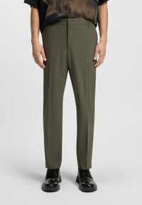 Olijfgroene, op maat gemaakte broek met een ontspannen pasvorm, voorzakken, riemlussen en een soepele textuur. Gecombineerd met zwarte veterschoenen.