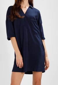 Donna che indossa un vestito camicia blu navy, largo, con maniche a tre quarti e una tasca sul petto, in piedi contro uno sfondo chiaro e uniforme.