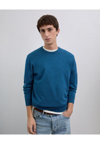 Niebieski sweter z dzianiny z okrągłym dekoltem i ściągaczami przy mankietach, zestawiony z białym t-shirtem i casualowymi dżinsami. Strukturę tkaniny widać jako miękką.