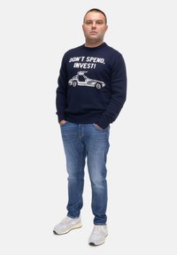 Maglione blu navy con grafica di una macchina bianca e testo "NON SPENDERE, INVESTI!" abbinato a pantaloni jeans azzurri chiari e scarpe sportive grigie.