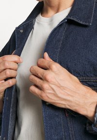 Ciemnoniebieska kurtka dżinsowa z kołnierzykiem, zapinana na guziki, z kontrastowym żółtym przeszyciem, noszona na jasnoszarej koszulce.