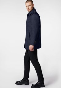 Granatowy wodoodporny płaszcz z wysokim kołnierzem, akcentami guzików i bocznymi kieszeniami, w połączeniu z dopasowanymi czarnymi spodniami i masywnymi czarnymi butami.