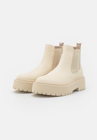 Bottines en cuir beige avec des panneaux latéraux élastiques, semelles épaisses et surélevées, et un contraste de surpiqûres. Texture lisse et bout rond.