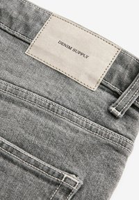 Graue Denim-Jeans mit einem hellgrauen Lederetikett, auf dem "DENIM SUPPLY" gedruckt ist. Auffällige Nähte und Taschen sind sichtbar.