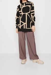 Osoba ubrana w czarną bluzkę z długim rękawem z beżowym abstrakcyjnym geometrycznym wzorem, fioletowe szerokie spodnie i beżowe platformowe loafersy.