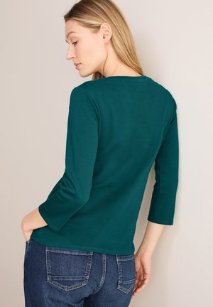 Türkises Oberteil mit drei Viertelärmeln, runder Ausschnitt, glatter Textur und lässiger Passform. Kombiniert mit dunkelblaue Jeans.