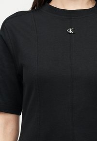 Μαύρο πουκάμισο με κοντά μανίκια, ανασηκωμένες κάθετες ραφές και μικρό λευκό έμβλημα με το λογότυπο CK κεντραρισμένο κοντά στη γραμμή του λαιμού.