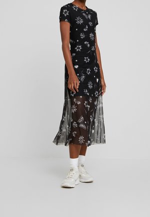 Persona che indossa un vestito midi nero trasparente con motivo floreale e di farfalle, calzini bianchi e sneakers bianchi su uno sfondo semplice.