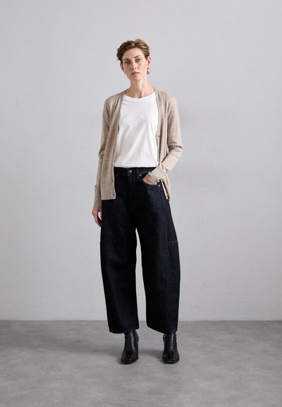 Beige cardigan over a white shirt, paired with dark denim wide-leg pants. Black ankle boots. Neutral background.
