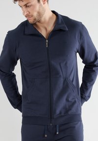 Navy zip-up jacket in soft cotton material, featuring a stand collar, side pockets, and ribbed cuffs. Subtle logo detail on the chest.