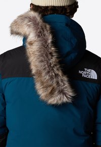 Blå parkas med en svart axelpanel, med en fuskpäls i ljusbrun vid huvan. Varumärkeslogotypen på den övre ryggen i vitt.