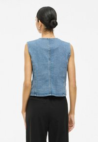 Denim prsluk bez rukava u svijetloplavoj boji, s prilagođenim leđima, vidljivim šavovima i blago skraćenim krojem. Glatka tekstura, minimalni dizajn.