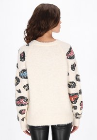 Kremowy sweter z cekinowym zwierzęcym wzorem na długich rękawach, teksturowanym dzianinowym wykończeniu i ściągaczami. Gładka tylna strona bez dodatkowych detali.