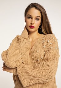 Cardigan tricotat bej, cu model împletit în cablu, având accente mari de perlă pe umeri și închidere cu nasturi.