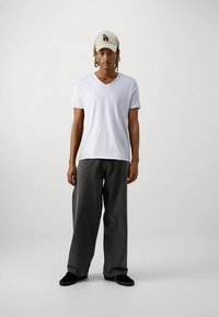 Biała koszulka z dekoltem w serek, luźne szare dżinsy i czarne buty wsuwane. Na głowie beżowa czapka z czarnym logo. Prosta postawa na neutralnym tle.