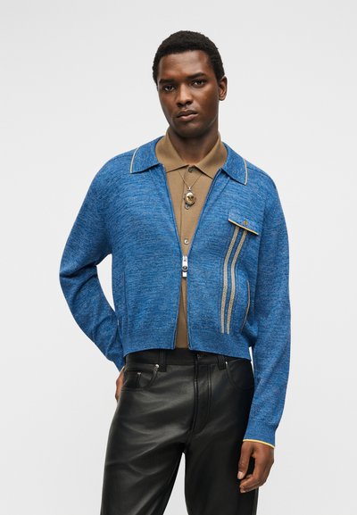 Άνδρας που φοράει μπλε ζακέτα με φερμουάρ πάνω από καφέ πόλο, μαύρο δερμάτινο παντελόνι και ένα στρογγυλό μενταγιόν στο λαιμό, ποζάρει μπροστά σε μονόχρωμο φόντο.