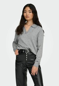 Grauer Strickpullover mit V-Ausschnitt und Kragen, kombiniert mit hochtaillierten schwarzen veganen Lederhosen mit Knopfdetails an der Vorderseite.