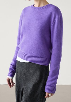 Femme portant un pull côtelé lavande à col rond manches longues sur une chemise blanche, associé à un pantalon taille haute gris foncé.